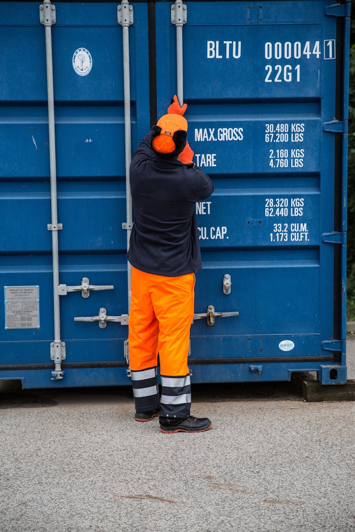 Naslovnica Industrial worker securing a shipping container outdoors, highlighting safety and logistics.