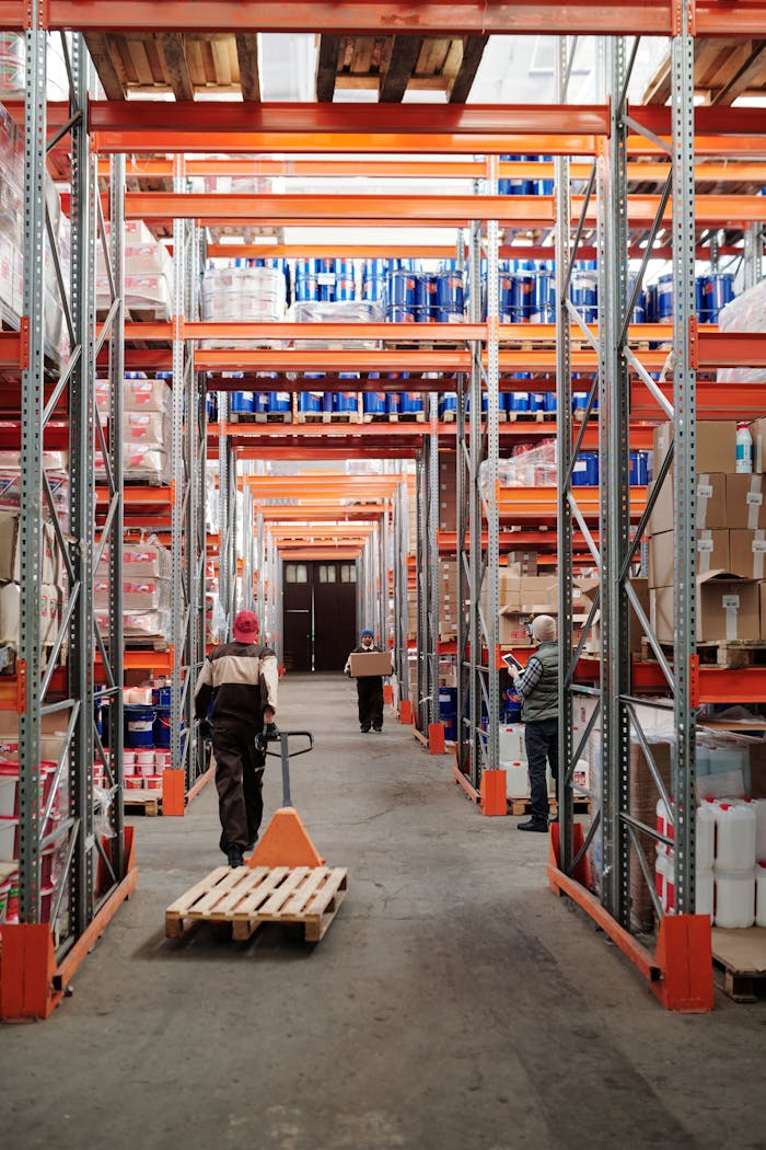 Naslovnica Workers arrange inventory on shelves in a large industrial warehouse.