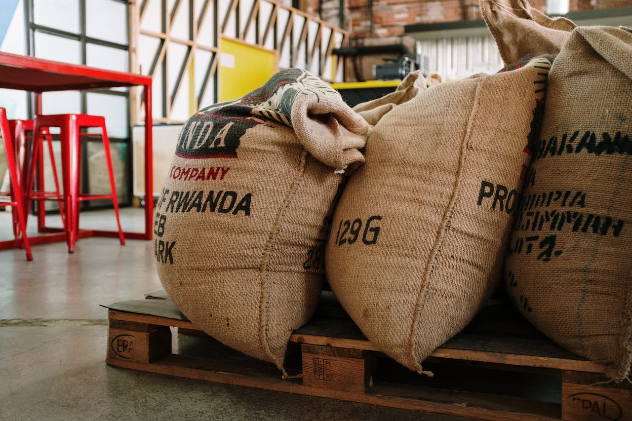 Naslovnica Burlap sacks of coffee beans stacked on a wooden pallet in an industrial warehouse setting.